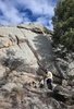 Person wearing outdoor clothing and a hat sitting on rocks at the base of a large granite boulder with trees and a partly cloudy sky in the background