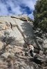 Man wearing an outdoors hat and jacket sitting on a large rock surrounded by boulders and trees under a blue sky with scattered clouds