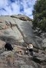 Man in outdoor gear sitting on rocks beside a small campfire near a large granite rock formation with a black and white dog sniffing nearby under a partly cloudy sky