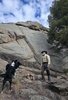 Man wearing a hat and hiking clothes standing on rocks near a small campfire, accompanied by two alert black and white border collies, with a large rocky cliff and pine trees in the background