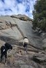 Person wearing a hat climbing large rocky terrain with two black and white dogs standing nearby on rocky ground under a partly cloudy sky