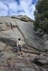 Person wearing a cowboy hat and beige jacket standing on rocky ground near a small campfire with a large rock formation and tree in the background under a partly cloudy sky