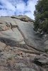 small campfire burning on rocky ground at the base of a large rock formation under a partly cloudy sky
