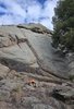 Small campfire burning at the base of a large rocky cliff under a partly cloudy sky with a tree on the right
