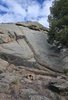 Large rugged rock formation with a small fire burning on the ground in front, surrounded by sparse vegetation and a tree on the right, under a partly cloudy sky
