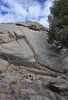 small campfire burning on rocky ground in front of a large rock formation with pine trees on the side under a partly cloudy sky