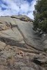 small fire burning on rocky ground in front of a large rock face with a tree to the right under a partly cloudy sky