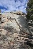 Large sunlit rock formation surrounded by pine trees under a partly cloudy sky with a black dog near the bottom right corner