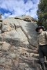 Older man in a tan jacket and black wide-brimmed hat standing near a campfire at the base of large rocky formations with pine trees and a partly cloudy blue sky overhead