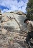 Elderly man wearing a wide-brimmed hat and tan jacket walking beside a large rock formation under a partly cloudy blue sky, surrounded by trees and dry grass