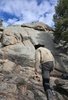 Person wearing a beige jacket and black hat crouching by a small fire at the base of large rock formations with trees nearby under a partly cloudy sky