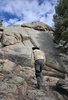 Person with a black hat, beige jacket, and dark pants climbing rocky terrain with pine trees nearby under a partly cloudy sky