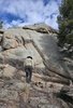 Person wearing a hat and jacket standing on rocky ground facing a large rock formation under a partly cloudy sky with trees nearby