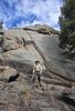 A man wearing a black cowboy hat and hiking clothes stands on rocky terrain beside a small fire, with a black and white border collie nearby and large boulders and trees around them