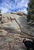 Large sunlit rock formation with scattered small plants at its base and a black dog with a bushy tail in the lower right corner, partially visible and standing on rocky ground