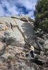 Person wearing outdoor clothing and a hat sitting on a rock at the base of a large rocky cliff with pine trees nearby under a partly cloudy sky