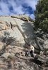 A person in outdoor clothing sitting on rocks near a large boulder with pine trees beside them under a blue sky with scattered clouds