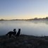 Two border collies standing on a frosty riverbank at dawn, looking out over a calm river with mist rising and a pale sky transitioning from night to morning