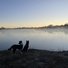 Two border collies standing on frosty ground by a calm river with mist rising and a pale sunrise in the background