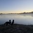 Two border collies standing on frosty grass at the edge of a calm river with mist rising and a clear sky at sunrise