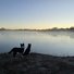 Two border collies stand on frosty grass by a calm river with morning mist rising and a clear sky at sunrise