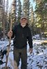 Older man with a white beard wearing a cap and black hoodie, holding a wooden walking stick, standing on a snowy forest trail with evergreen trees and a black and white dog behind him