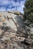 Large sunlit rock formation with a small campfire in front and evergreen trees around, under a partly cloudy blue sky