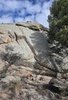 Large rocky cliff face with a small fire burning at its base and pine trees partially shading the area under a blue sky with scattered clouds