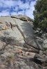 Small campfire burning at the base of a large rock formation with pine trees and a partly cloudy sky above
