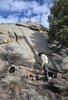 Person wearing a hat and beige jacket tending a small campfire among large rocky boulders with two black and white dogs nearby under a partly cloudy sky