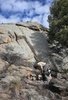 person bending over near a small campfire on rocky terrain with a black and white dog standing nearby and large rock formations and a pine tree in the background under a partly cloudy sky