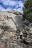 Person in outdoor clothing sitting on rocks near a small campfire at the base of a large rock formation with trees nearby under a partly cloudy sky
