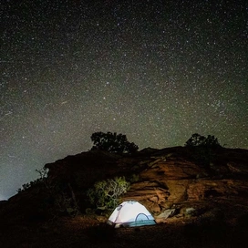 A lit camping tent set among desert rocks under a clear night sky filled with countless stars