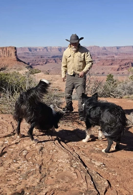 Man wearing a hat and beige jacket standing on rocky terrain with two black and white dogs, with layered red rock cliffs and canyons under a clear blue sky in the background