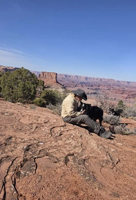 A person wearing a wide-brimmed hat and tan jacket sitting on red rock terrain, petting a black and white dog, with another dog standing nearby and expansive canyon cliffs in the background under a clear blue sky