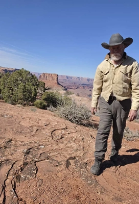 Older man with a white beard wearing a wide-brimmed hat and beige jacket walking on red rocky terrain with shrubs and a cliff formation in the background under a clear blue sky