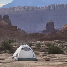 A solitary gray and white tent set up on flat rocky terrain with sparse desert bushes, surrounded by towering layered red rock formations under a hazy sky