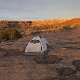A small tent set up on a flat rock surface surrounded by sparse desert vegetation and rocky cliffs bathed in warm, orange sunlight