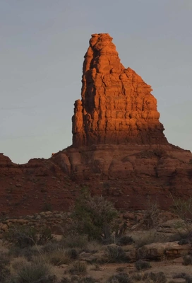 Tall, vertical red sandstone rock formation illuminated by warm sunlight against a pale sky, with desert vegetation and rocky terrain in the foreground