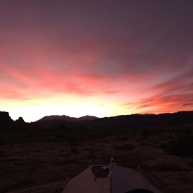 vibrant sunset sky with shades of orange, pink, and purple over a rocky desert landscape with silhouetted cliffs and a tent in the foreground