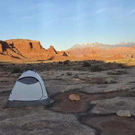 Small tent set up on rocky desert terrain with towering red rock cliffs and distant snow-capped mountains under a clear blue sky
