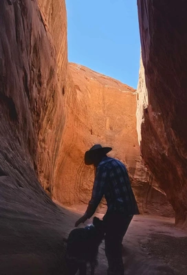 Person wearing a wide-brimmed hat and plaid shirt standing inside a narrow canyon with smooth red rock walls and a dog