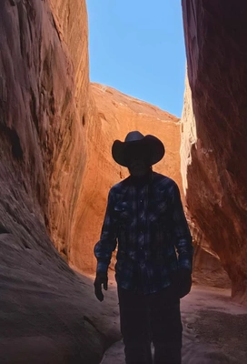 Person wearing a cowboy hat and plaid shirt standing in a narrow sunlit canyon with tall red rock walls and a clear blue sky above