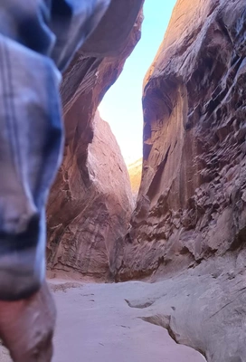 Narrow, towering red rock canyon walls illuminated by sunlight, with a sandy path winding through the middle and part of a person wearing a blue plaid shirt visible on the left side