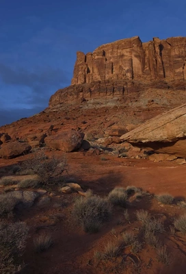massive towering red sandstone cliffs under a dark blue sky, with a rocky desert foreground and sparse desert vegetation