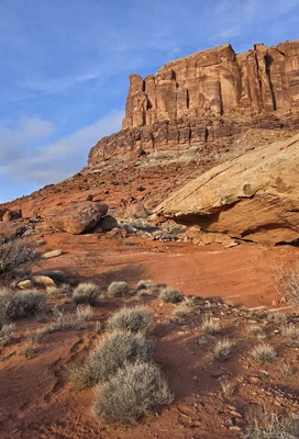 Red rock canyon landscape with a layered sandstone cliff under a partly cloudy blue sky and desert shrubs in the foreground