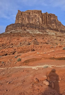 Large reddish-brown sedimentary rock formation towering over rugged red sandstone landscape with sparse desert vegetation under a partly cloudy blue sky