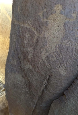 ancient petroglyphs carved into a rock face, depicting human-like and animal-like figures with raised arms and various abstract shapes