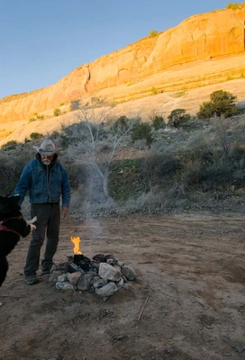 Man wearing a hat and denim jacket interacting with a dog beside a small campfire with orange rocky cliffs in the background during daylight