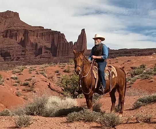 Man wearing a cowboy hat riding a horse in a red rock desert landscape with tall rock formations in the background